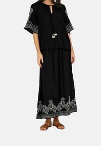 Conjunto negro que incluye una blusa holgada con detalles de borlas y bordados decorativos en blanco, combinada con una falda larga a juego y sandalias marrones.
