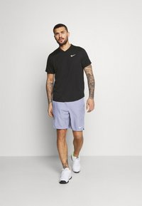 Camiseta deportiva negra con cuello en V y logo blanco, combinada con pantalones cortos lilas claros con un logo. Zapatillas deportivas blancas con detalles verdes.