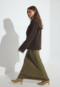 Hnedá semišová bunda, olivovo zelená dlhá sukňa, béžové špicaté podpätky. Textúrovaná látka s viditeľnými švami a jednoduchou siluetou.