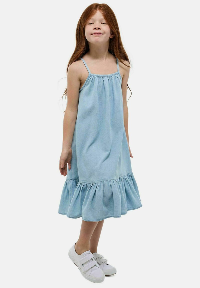 Robe en denim bleu clair avec des bretelles spaghetti, un corsage froncé et un ourlet festonné. Associée à des baskets blanches. Texture lisse, design décontracté.