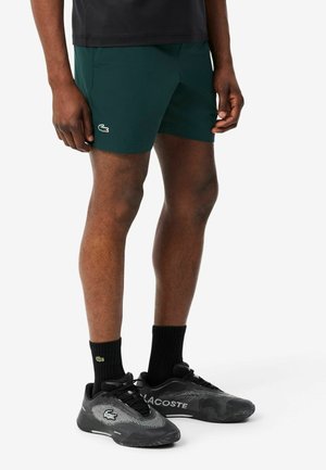 Uomo che indossa pantaloncini verde scuro con un piccolo logo di coccodrillo, calzini neri con logo e sneakers Lacoste nere su sfondo bianco.