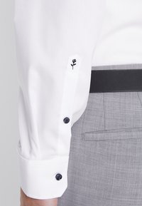 Ärmels eines weißen Hemd mit einer schwarzen bestickten Rose nahe der Manschette, schwarze Knöpfe, kombiniert mit grauer Hose und einem schwarzen Gürtel.