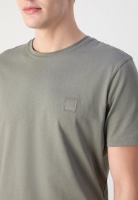 Homme portant un T-shirt olive à manches courtes avec un petit patch logo carré "BOSS" sur le côté gauche de la poitrine, recadré de la bouche au milieu du torse.