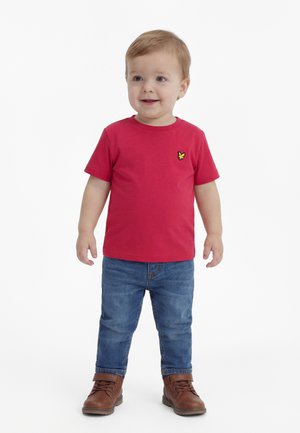 Tout-petit aux cheveux châtain clair portant un t-shirt rouge avec un logo jaune, un jean bleu et des bottes marron, debout et souriant sur un fond blanc.