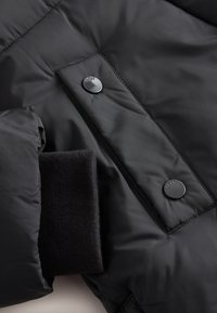 Veste matelassée noire dotée d'un tissu extérieur lisse, de boutons-pression et d'un détail de poignet noir texturé. Idéale pour les temps froids.