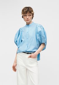 Γυναίκα που φοράει ανοιχτό μπλε μπλούζα με φουσκωτά μανίκια με οπές και άσπρο παντελόνι, στέκεται με το ένα χέρι στην τσέπη σε ένα απλό φόντο.