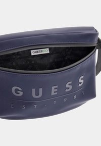 Borsa in vita sintetica blu navy con texture e logo "GUESS" in rilievo. L'apertura con zip rivela un interno a motivo. Design durevole e compatto.