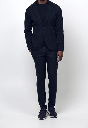 Homme portant un blazer noir, un t-shirt noir, un pantalon slim noir et des baskets noires sur un fond clair uniforme.
