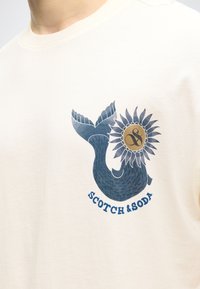 Κοντινή λήψη κρεμ t-shirt με μπλε ουρά φάλαινας και γραφικό ηλίανθου πάνω από το κείμενο «Scotch & Soda» στο στήθος.