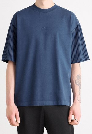 Personne portant un T-shirt BOSS bleu marine à manches courtes rayées et un pantalon noir, montrant un tatouage de serpent sur l'avant-bras gauche.