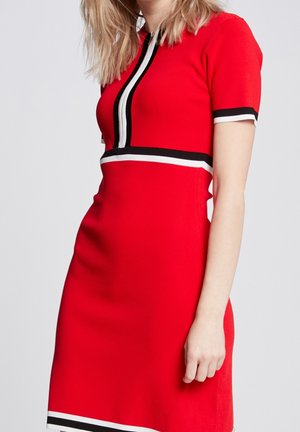 Kobieta w dopasowanej czerwonej sukience z krótkimi rękawami i czarno-białymi pionowymi oraz poziomymi paskami na piersi i rękawach.