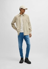 Béžová kordová košeľa s gombíkmi, vrstvená na bielom tričku, v kombinácii s modrými džínsami a čiernymi robustnými čižmami, doplnená béžovou čiapkou.