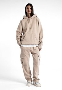 Hoodie en polaire beige avec une poche avant et un cordon de serrage, associé à un pantalon cargo assorti avec des poches latérales et une coupe décontractée. Des baskets terminent le look.