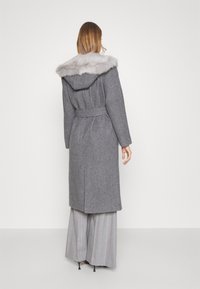 Woman wearing a belted gray wool coat with a fur-trimmed hood, wide-legged pinstripe trousers, and black high heels, standing facing away.