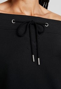 Felpa nera off-the-shoulder con texture a costine, presenta una chiusura con cordino e dettagli in argento, insieme a una sottile cucitura decorativa.