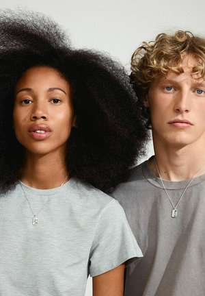 Due persone stanno vicine, indossando t-shirt casual—una grigia chiara, l'altra grigia scura. Entrambe indossano collane d'argento con pendenti rettangolari.