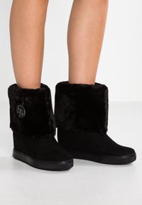 Botas de cuña negras hasta la mitad de la pantorrilla con gruesos puños de piel sintética y un emblema metálico redondo en el lado exterior, llevadas sobre piernas desnudas.