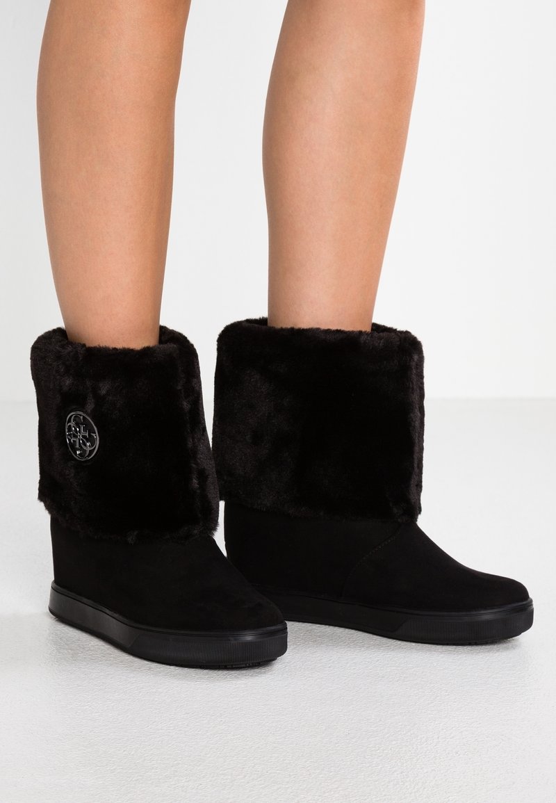 Botas de cuña negras hasta la mitad de la pantorrilla con gruesos puños de piel sintética y un emblema metálico redondo en el lado exterior, llevadas sobre piernas desnudas.