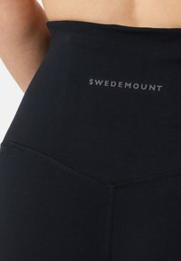 Leggings noirs taille haute en tissu extensible avec une texture lisse, arborant un logo "SWEDEMOUNT" en gris au niveau de la taille.