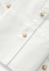 Hvid bomuldsstof med en glat tekstur, prydet med lysebeige knapper og præcise sydetaljer. Nærbillede af stoffet og lukkemekanismer.