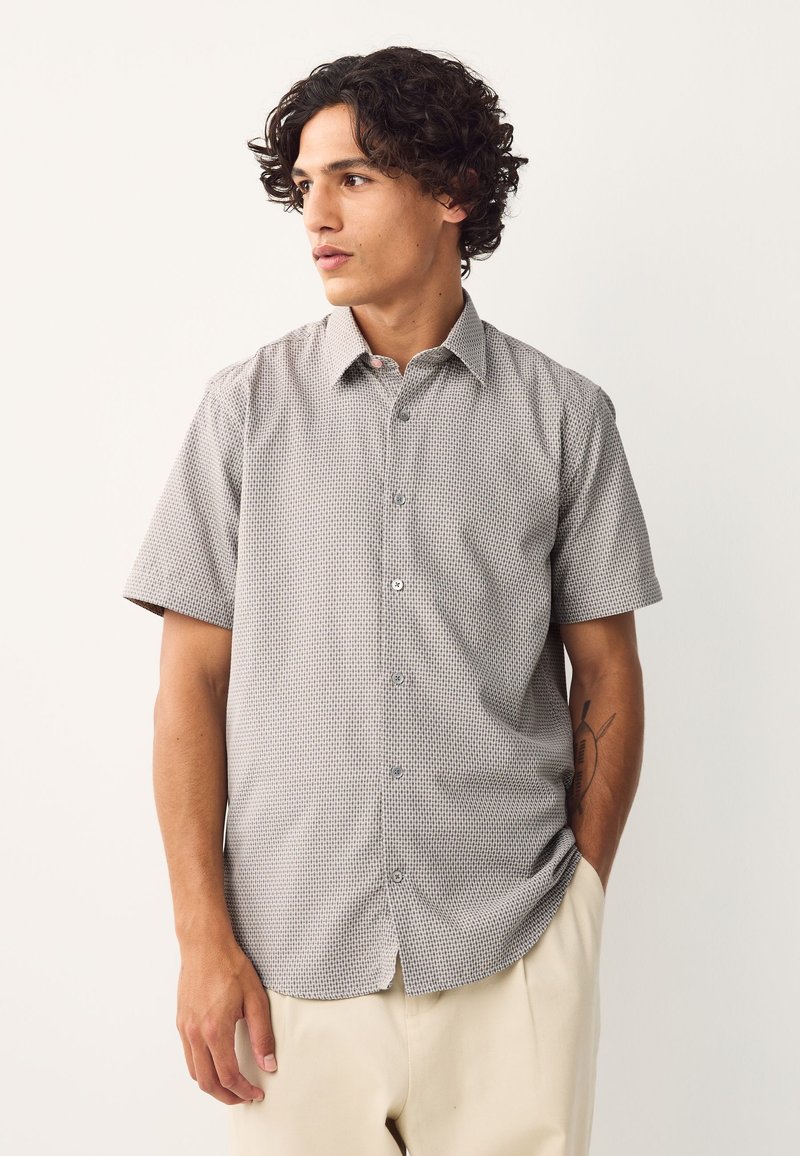 Jeune homme aux cheveux bouclés portant une chemise à manches courtes à boutons avec un motif et un pantalon clair, debout avec une main dans la poche.