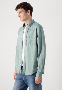 Lys grønn, langermet skjorte med krage som kan kneppes, frontlomme og hvite knapper, brukt over en hvit T-skjorte og blå jeans.
