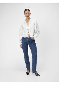 Camicetta bianca corta con scollo arricciato e maniche in pizzo, abbinata a jeans blue dritti e décolleté neri con punta. 