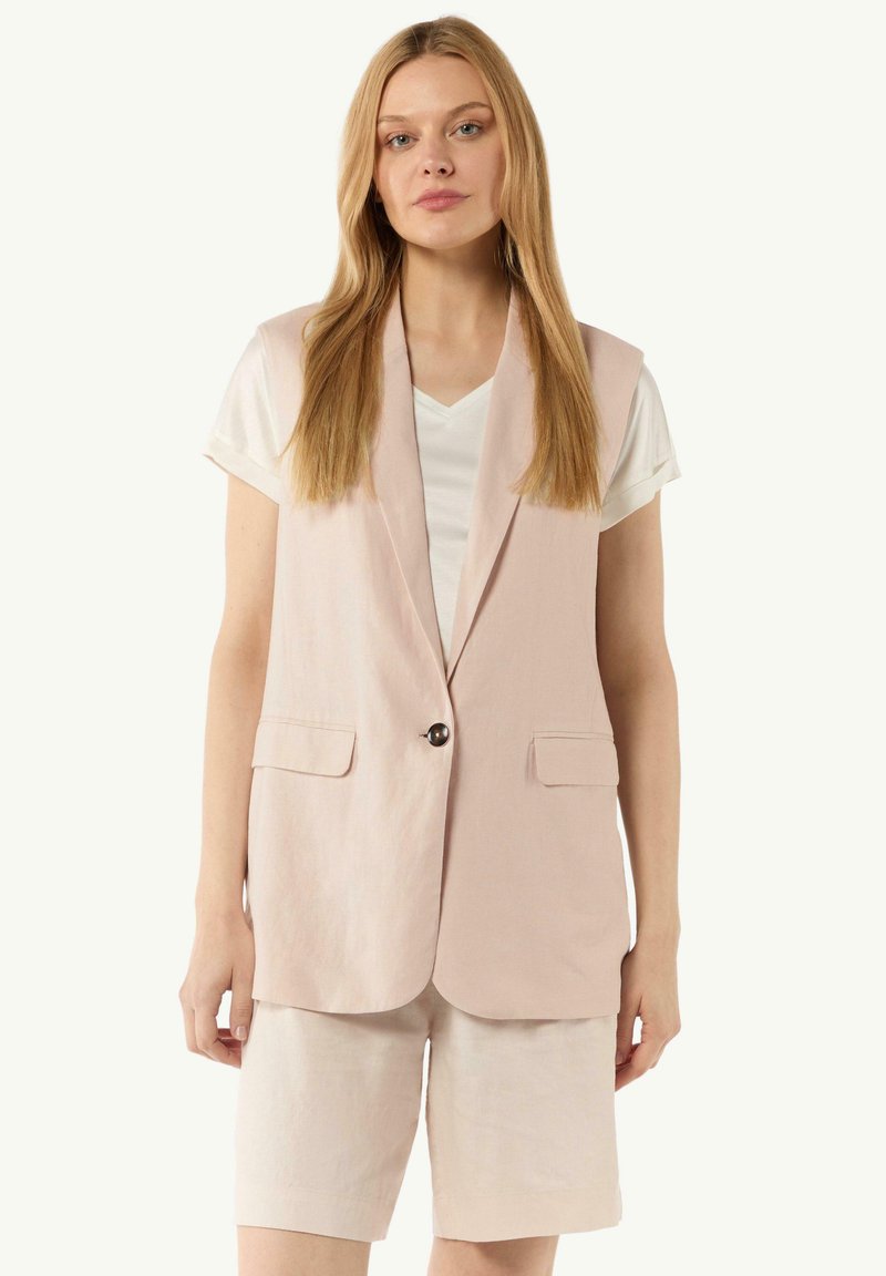 Gilet en lin rose avec une fermeture à un seul bouton, comportant deux poches avant, porté sur une chemise blanche à col en V et associé à un short beige.