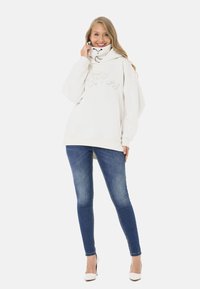 White oversized sweatshirt with a high collar and embroidered "Legendary" text, paired with dark blue skinny jeans and white heels.