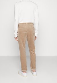 Pantalon en velours côtelé beige à coupe droite, avec une ceinture munie de passants, deux poches arrière, et des revers au-dessus de baskets blanches.