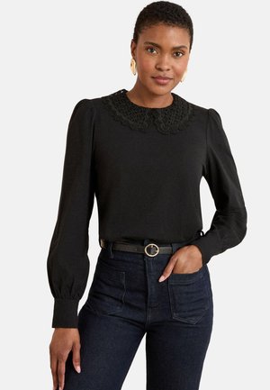 Frau mit einer schwarzen Bluse mit Spitzenkragen, langen Puffärmeln, in dunklen High-Waist-Jeans mit einem schwarzen Gürtel mit runder Schnalle gesteckt.