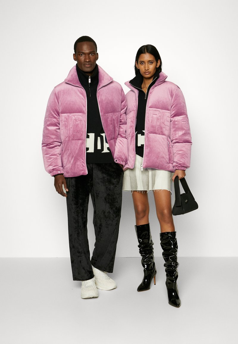 Due modelli che indossano giacche a vento in velluto rosa abbinate; uno con pantaloni neri e scarpe bianche, l'altro con una gonna leggera, stivali neri e in mano una borsa.