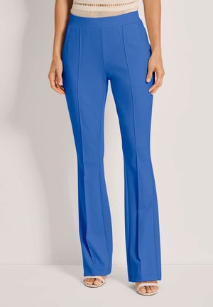 Frau trägt eine hochtaillierte, hellblaue, gerade geschnittene Hose, kombiniert mit weißen Sandalen mit offenen Spitzen und einem beigen Oberteil.