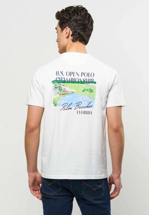 Mężczyzna noszący białą koszulkę z grafiką U.S. Open Polo Championship Palm Beaches Florida i ilustracją krajobrazu wybrzeża na plecach.