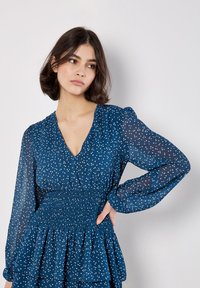 Marineblaues Kleid mit weißen Polka-Dots, V-Ausschnitt, geraffter Taille und langen, durchsichtigen Ärmeln. Es hat einen mehrlagigen Rüschensaum am Rock.