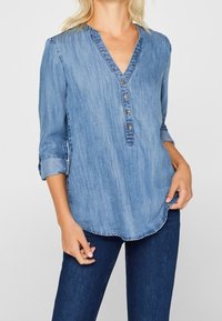Jasnoniebieska koszula dżinsowa z dekoltem w serek, zapięciem na guziki i rękawami o długości trzech kwart. Miękka textura i zaokrąglony krój.