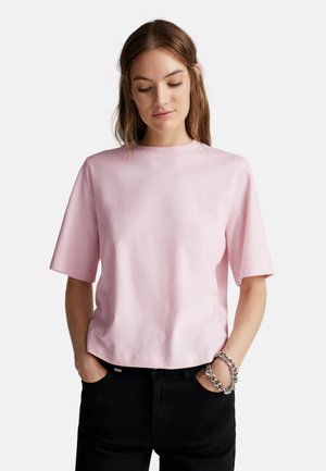 Jonge vrouw met bruin haar, gekleed in een lichtroze shirt met korte mouwen, zwarte broek en een stevige zilveren armband, met handen in de zakken en omlaag kijkend.