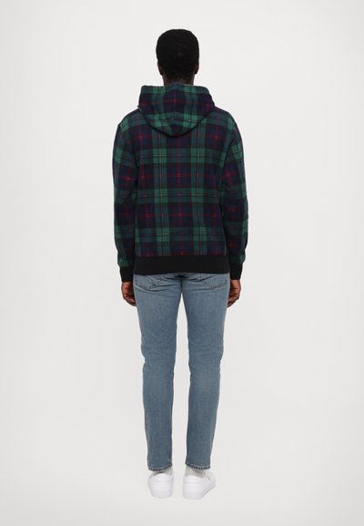 Felpa a quadri verde e navy con accenti rossi, dotata di cappuccio con coulisse e polsini a costine, abbinata a jeans azzurri e sneaker bianche.
