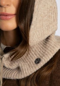 Beige gebreide capuchon met geribbelde textuur, voorzien van een donkere decoratieve knoop. Zacht materiaal valt rond het halsgebied.