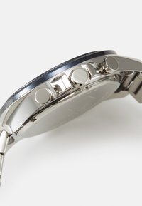 Un reloj de acero inoxidable con un bisel azul, que presenta botones de presión, una corona texturizada y una superficie suave y reflectante.