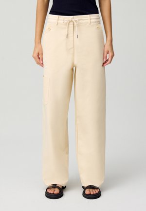 Pantalones de pierna ancha color beige, hechos de una tela suave, con cintura ajustable, bolsillo lateral y dos botones frontales. Textura lisa y diseño relajado.