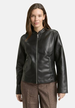 Frau trägt eine schwarze Lederjacke mit Reißverschluss und zwei Reißverschlusstaschen sowie braune Hose, steht vor einfarbigem Hintergrund.