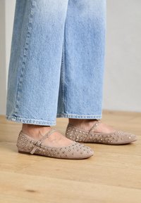 Beige mokasiner i semsket skinn med rund tå, prydet med sølvspiker og justerbar ankelrem, parret med lyseblå vide jeans.