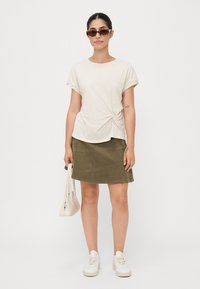 Licht beige knoopvoor t-shirt, olijf groene suede mini rok, witte sneakers en een kleine witte handtas, gecombineerd met donkere zonnebril.