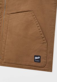 Braune Canvas-Jacke mit einem großen Fronttasche und einem schwarzen Etikett mit weißem Text. Glatte Textur mit sichtbaren Nähten.