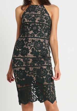 Femme portant une robe noire sans manches en dentelle florale sur un jupon couleur chair, debout devant un fond clair uni.