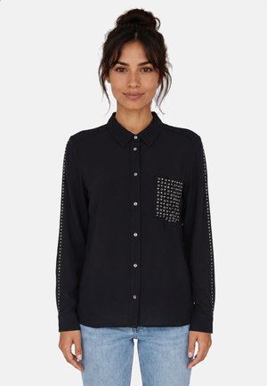 Donna con capelli scuri raccolti in uno chignon che indossa una camicia nera con bottoni e borchie d'argento sulla tasca sul petto e sulle maniche, abbinata a jeans azzurri chiari.