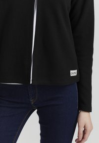 Černá ribbed bunda s otevřeným předním dílem, se bílou etiketou na lemu, v kombinaci s tmavě modrými džíny. Hladká textura, ležérní design.