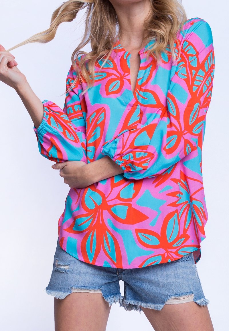 Haut coloré présentant un motif floral vibrant en rouge, rose et turquoise, avec de longues manches bouffantes et une coupe décontractée. Associé à un short en jean.