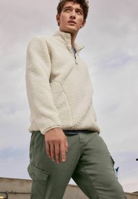 Pull en polaire crème avec un zip quart et une texture douce, associé à un pantalon cargo vert olive avec des poches latérales. Fond nuageux.